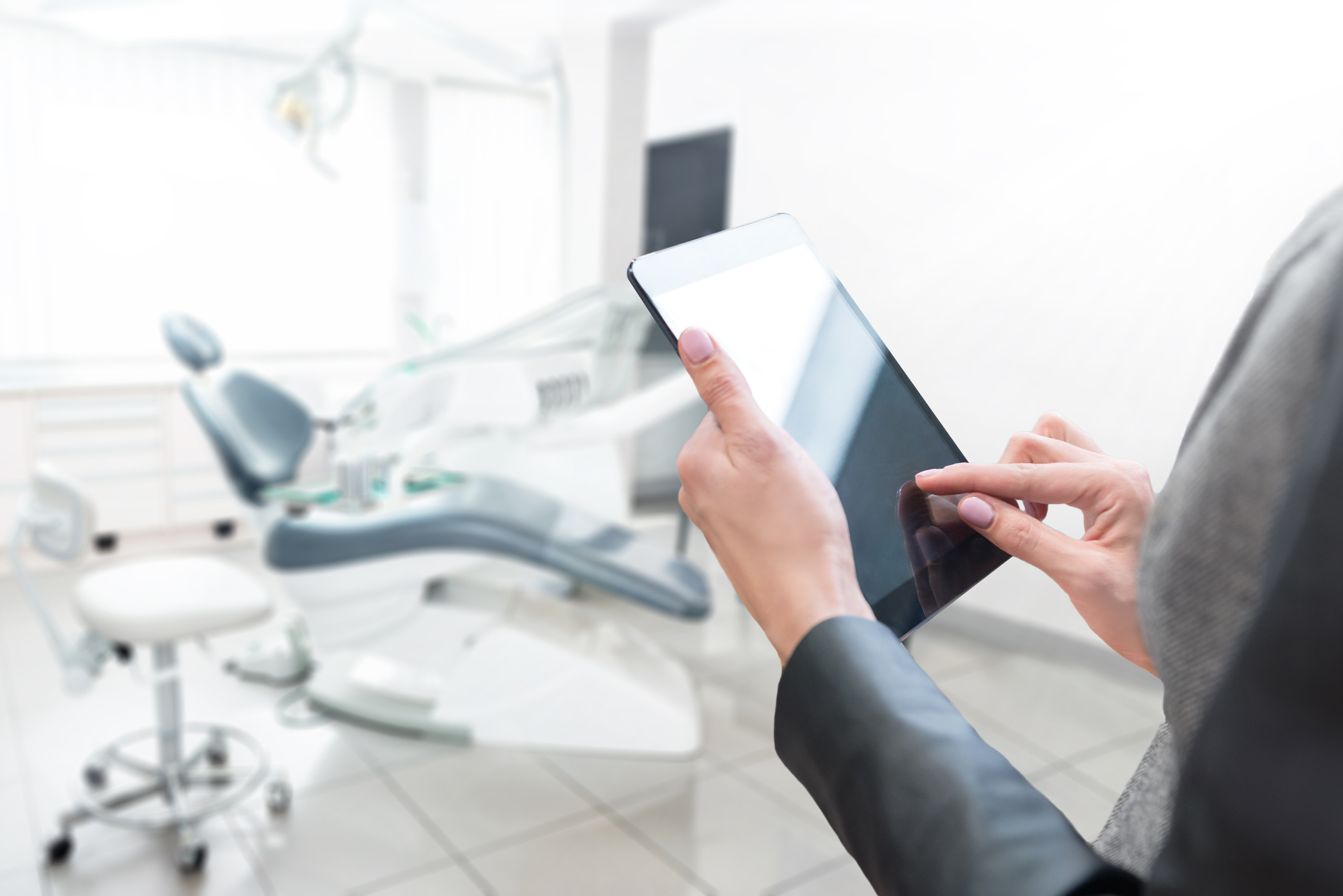 A woman working on a tablet in a dental office with a dental chair and equipment.